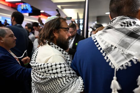 Seen closely from behind, a man with long hair and a beard wearing a yarmulke links arms with someone in front and behind him, all wearing keffiyehs.