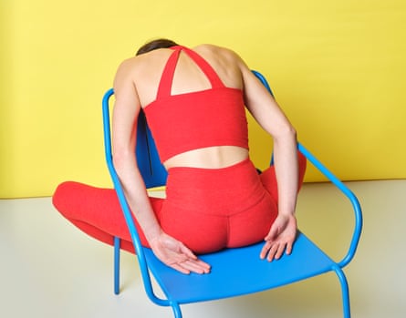 A woman in red gym gear in a relaxed pose leaning over the back of a blue metal chair against a yellow backdrop