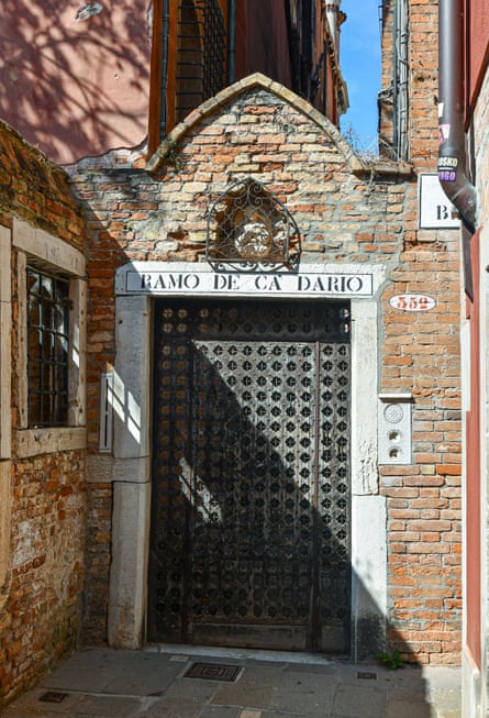 Arched brick and stone entrance gate to the palace with carved wooden gate and a barred window to the side of an alley; an inscription in black on white stone over the door reads Ramo De Ca’ Dario.