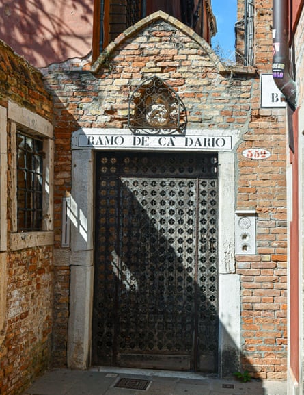 Arched brick and stone entrance gate to the palace with carved wooden gate and a barred window to the side of an alley; an inscription in black on white stone over the door reads Ramo De Ca’ Dario.