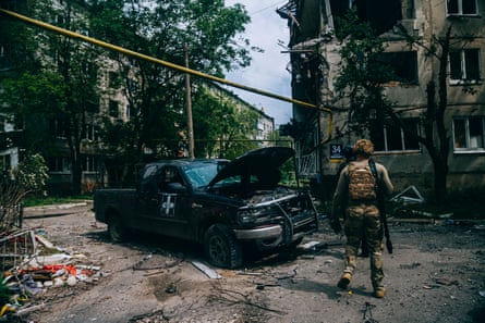 A person in military unifor near a burnt-out vehicle in an urban area badly damaged by fighting