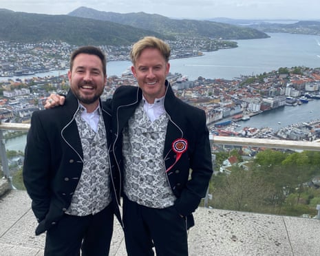 Martin Compston and Phil MacHugh, both with matching fancy jackets and waistcoats and both grinning, stand on a walkway with houses, mountains and a river behind them