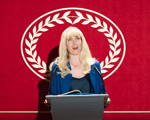 Kate O'Flynn, in character, speaks at a podium with two microphones