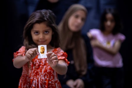 Aysel holds up a photograph of her father, with her mother and a brother in the darkened, blurred background. She is smiling and wears a red dress.