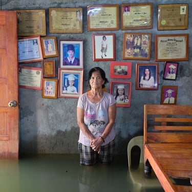 A woman stands in a house thigh-deep in flood water next to a wall covered in graduation pictures and certificates