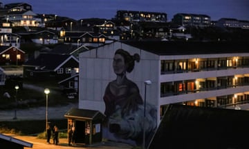 A mural of a woman and a polar bear on the side of a block of flats in an urban area lit up after dusk