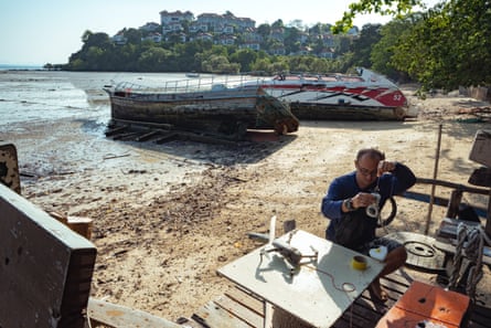 A man on the beach of a small bay squats on a platform with a camera in his hands. A small drone lies on the table beside him. Two boats pulled out of the water are behind him