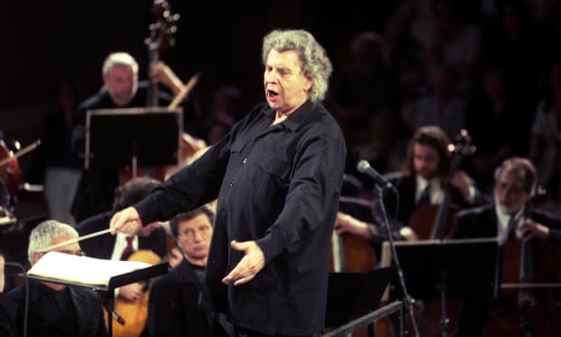 Theodorakis dressed all in black, his eyes closed and both arms low and outstretched with a conductor's baton in one hand, surrounded by orchestral musicians