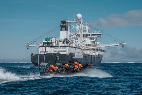A small boat full of people dressed in orange goes past a large fishing trawler on the sea
