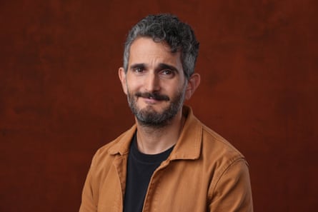 A man in a tan jacket and a black T shirt, with a grey beard