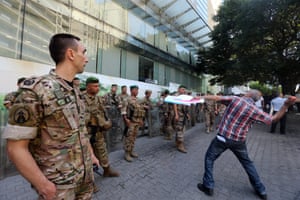 A man in civilian clothing throws a tomato, watched by a line of military personnel in camouflage gear