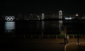 Two men in face masks walk alongside water in Tokyo, with Olympic rings behind them.