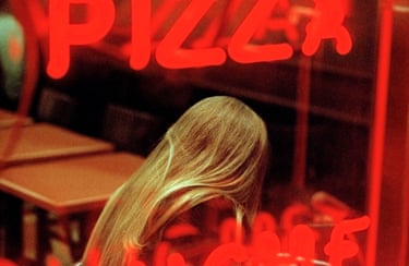 A young woman’s long blonde hair falls forward beneath a red neon “Pizza” sign