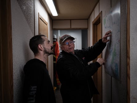Andy Rae, with dark beard and moustache, stands with an older man looking at a map on a wall in the narrow hallway of a building. The older man holds his hands to the map.