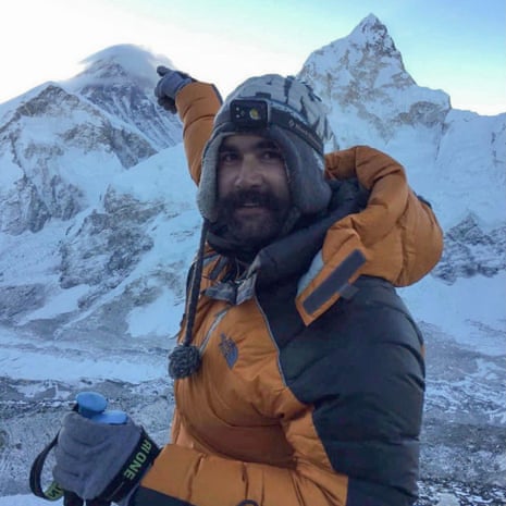 A man standing near the top of a mountain points upward, smiling, with snowy mountain peaks behind him