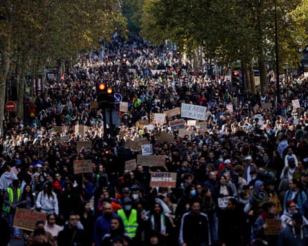 A dense crowd fills a Paris street during a protest, many holding handwritten cardboard signs