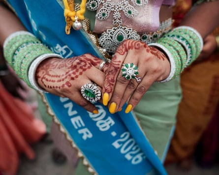 Closeup of a woman’s hands clasped in front of her pageant winner’s sash. She is wearing green glass bangles and jewellery, yellow nail polish and has henna on her hands