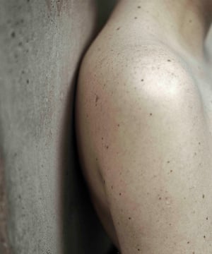 A bare shoulder, with all its moles and freckles, against a concrete wall