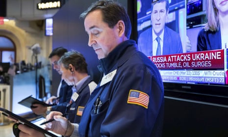 A man at the New York Stock Exchange uses a stylus on a tablet computer while a screen behind him broadcasts a news station with the caption "Russia attacks Ukraine: Stocks tumble, oil surges."