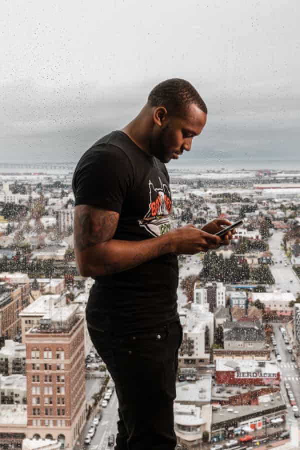 A man in a black shirt looks down at his phone, a cityscape seen through a window behind him