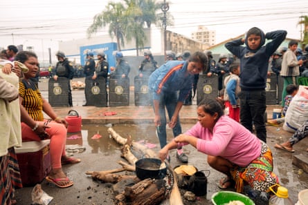 Seorang wanita mengurus api memasak yang dikelilingi oleh orang lain, sementara polisi anti huru-hara dapat terlihat di latar belakang.