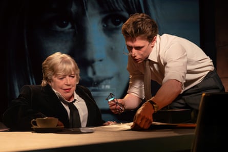 A woman in a black suit and tie sits at a desk while a young man leans over the desk showing her some documents.