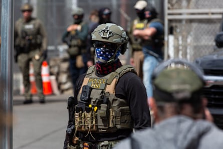 A person wearing a soldier's helmet and kevlar vest with an American flag bandana covering his mouth stood near to a metal fence