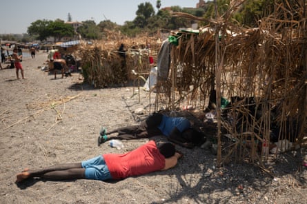 Several people on a beach lie in the shade on a sunny day underneath makeshift shelters.