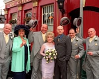 A lineup of wedding guests with bride Barbara Windsor and groom Mike Reid at centre outside the red painted Queen Vic pub - part of the pub title showing at top left of image