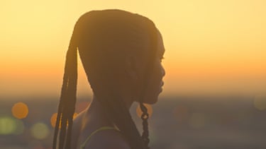A silhouetted woman with long braided hair gazes into the distance against a glowing sunset sky