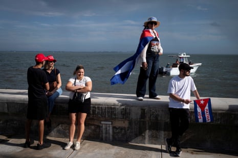 Protesters in red Trump hats and carrying Cuban flags stand against a parapet overlooking the ocean.