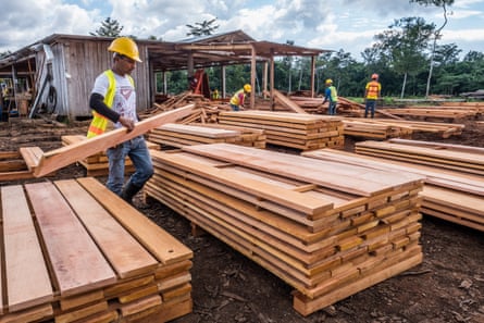 A man in a yellow hard hat moves a plank of wood on to one of several stacks of processed wood at a sawmill.