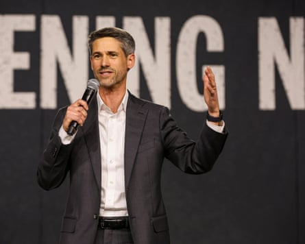 man in suit speaks on stage into microphone with hand raised