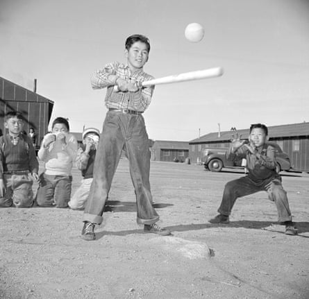 A boy swings a baseball bat at Manzanar as a ball flies past, children watching nearby