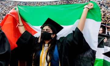 Young Middle Eastern woman wearing black mask and graduation robe and gown holds red, green and white Palestinian flag over her shoulders.
