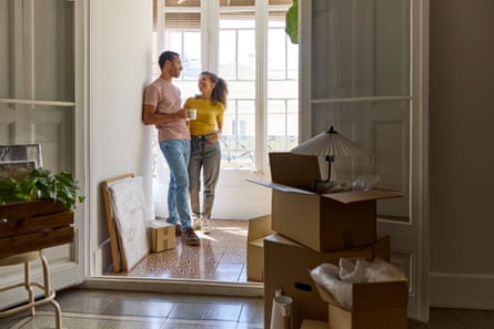 Smiling black or mixed-race youngish man and woman talking in a doorway in front of high windows; there are cardboard boxes in the foreground containing lamps and wrapped-up items which suggest they have just moved in to a property; the room’s woodwork is white and the floor tiled; they wear blue jeans and trainers and he wears a light pinkish T-shirt while hers is yellow; she has long, curly dark hair in a ponytail.