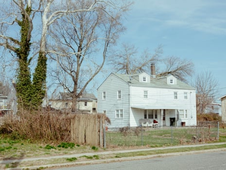 A scene of a few two-story homes in good condition, with large, flat, grassy yards, a few spare trees, and a thin sidewalk along a grassy verge.