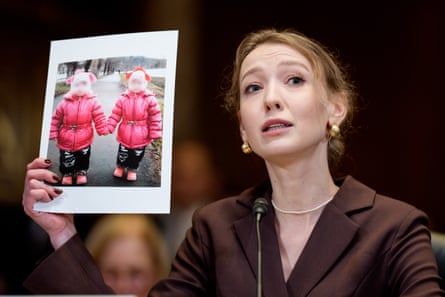 A young woman with a mic in front of her holds up a photo of two small children in pink jackets