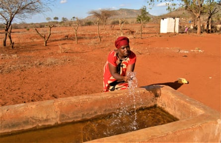 A woman in a dress and headscarf splashes water out of a large drinking trough in an expanse of terracotta-coloured ground