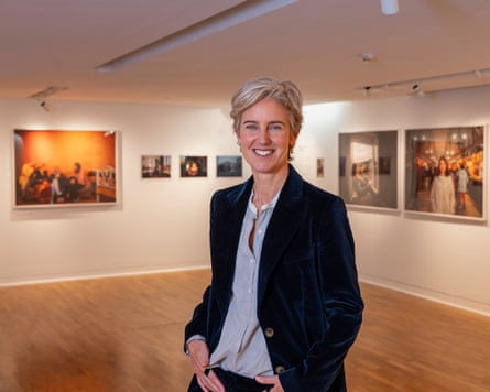 Photographer Polly Braden, in black jacket and collarless shirt, in a gallery with photos on the wall.