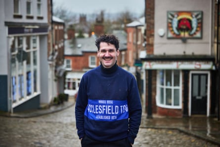 Tom Broadhead smiles as he stands wearing a jumper embroidered with the words ‘Macclesfield FC’