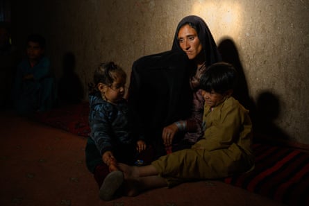 A sad-looking woman sits with two children in a dark room, with light from a window falling on the wall behind her.
