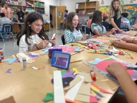 Children sit across from each other at a long line of desks with colored construction paper.