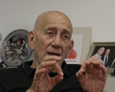 Ehud Olmert during an interview with The Guardian: he holds his hands up in an animated gesture. He is nearly 80, bald, and wears a black top; he sits in front of a shelf with photographs and an ornamental plate.