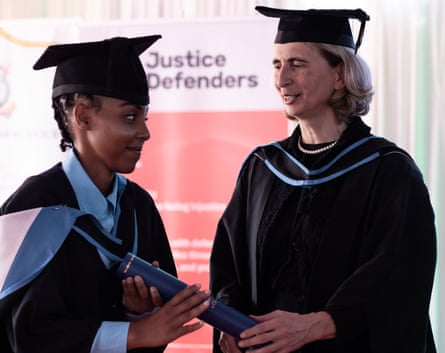Two women wearing graduation mortarboards and gowns stand in front of a poster for Justice Defenders
