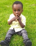 Awaab Ishak – two-year-old boy looking wide-eyed at the camera with his fingers in his mouth; he is sitting on grass and wearing a light shirt and grey trousers