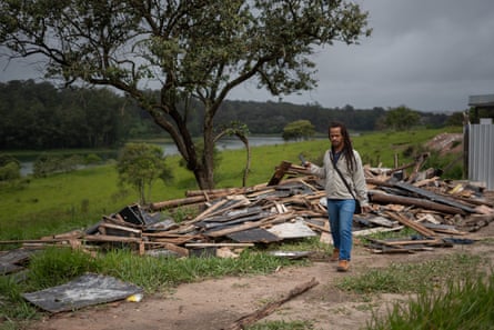 A man walks past a large pile of building materials dumped on grass under a tree near a body of water.