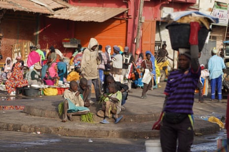 Four young boys sit on a small roundabout as another stands looking down at them, amid the bustle of a street market, with porters and women seated selling goods from big bowls