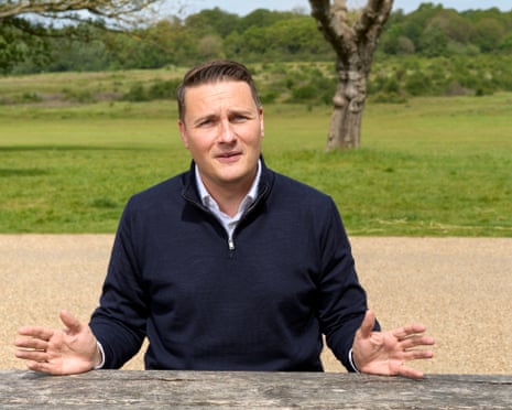 Wes Streeting in a navy jumper sits at a wooden table outdoors, gesturing with both hands raised