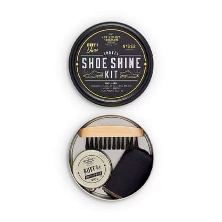 A Gentlemen’s Hardware Travel Shoe Shine Tin is open. At the top is the black lid with white text and on the bottom is the inside of the tin with a brush and a pot of buff. It is on a white background